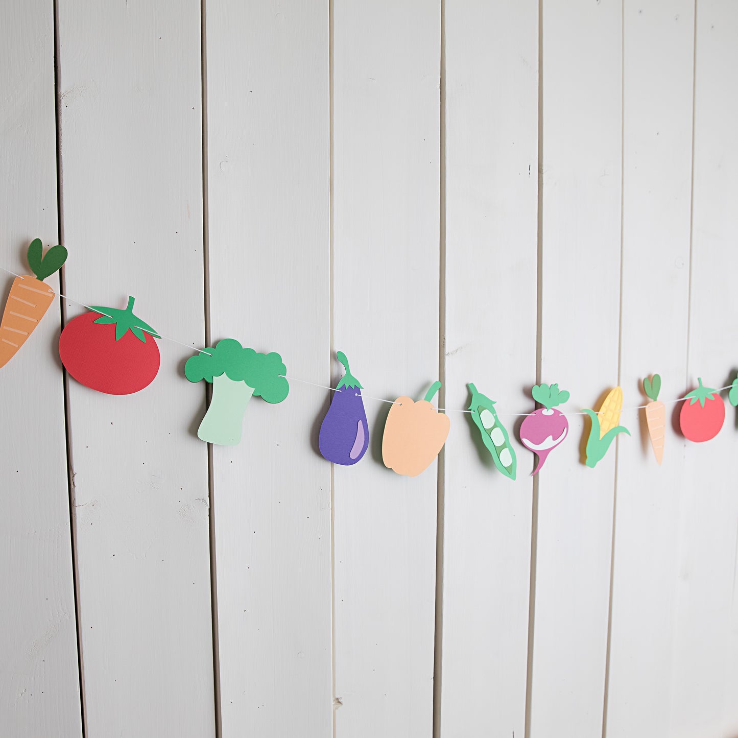 Colorful paper cutout vegetables hanging on a string against a white wooden background