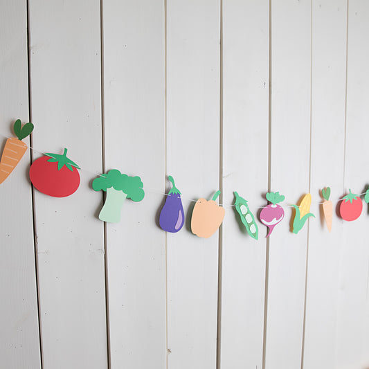 Colorful paper cutout vegetables hanging on a string against a white wooden background