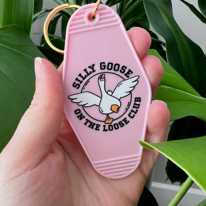Pink keychain with 'Silly Goose on the Loose Club' design held in a hand with green leaves in the background