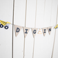A happy birthday banner with the word 'DIG IN' written on pennants attached to black twine, decorated with construction vehicle figures and placed against a white background.