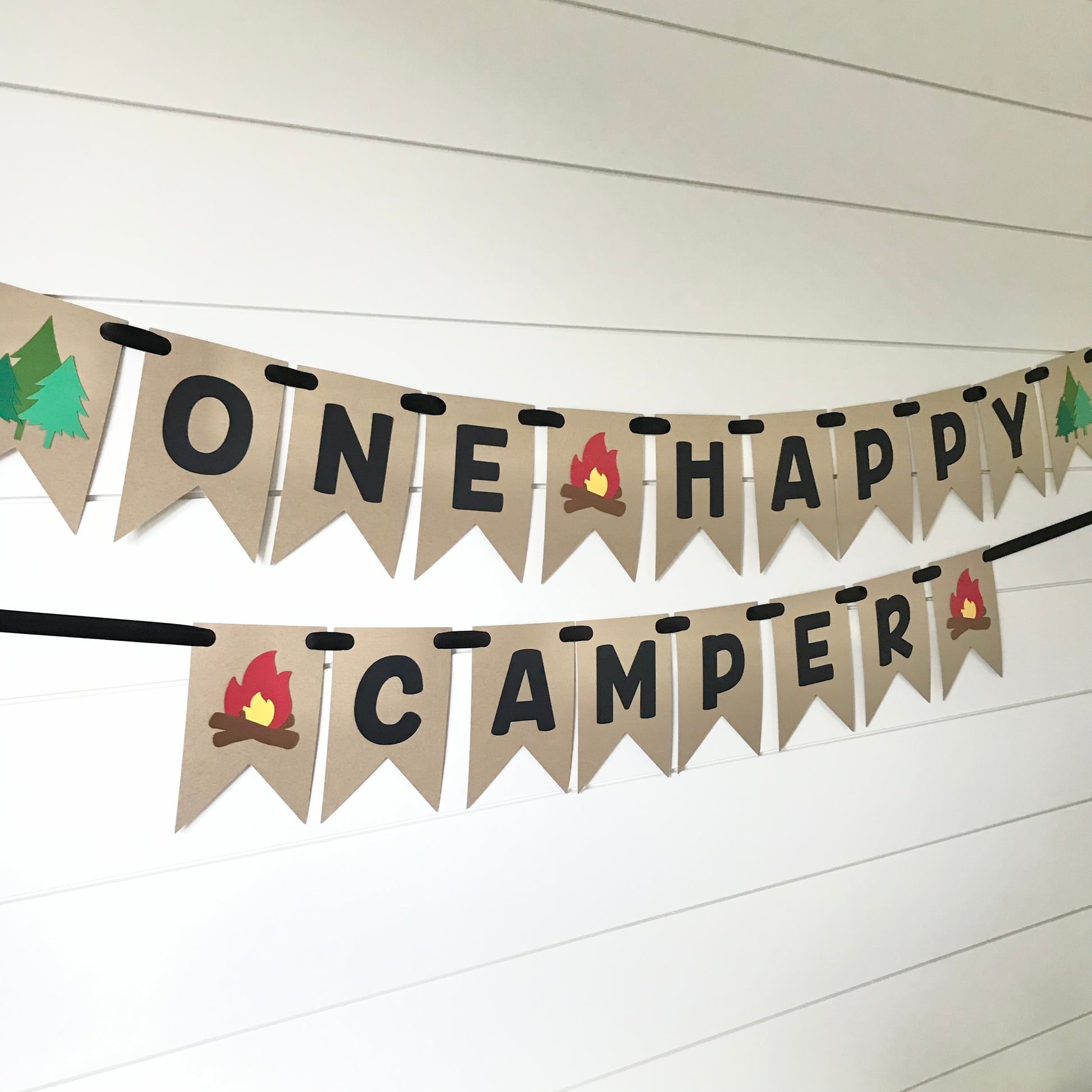 banner in kraft color that says "one happy camper" in black font with tree's and a fire on the ends
