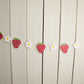 paper strawberries and daisies strung on twine against a wall 