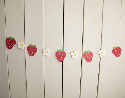 paper strawberries and daisies strung on twine against a wall 